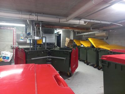 Garbage Doctor Duo in a long bin room with rows of red- and yellow-lidded waste bins