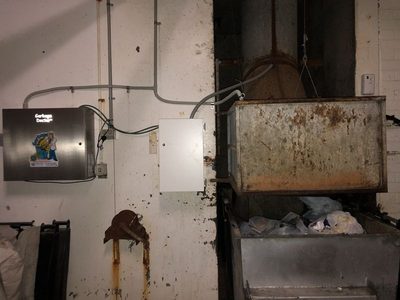 Garbage Doctor Duo and its scent tank beside a rusted chute hopper over a full galvanised rubbish bin