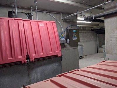 Garbage Doctor Duo beside a chute over large bins with red lids, at the edge of a car park