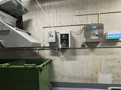 Garbage Doctor Duo and its scent tank on a concrete bin room wall, beside the chute's hopper and a green bin