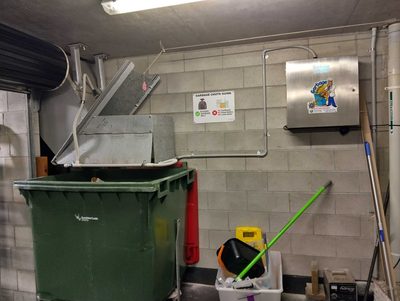 Garbage Doctor Ozone on a block wall beside the chute's hopper, over a green waste bin