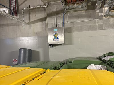 Garbage Doctor Ozone on a block wall above rows of yellow- and green-lidded recycling and waste bins