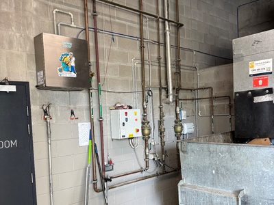 Garbage Doctor Ozone on the wall of a compactor room, above its pipes and a galvanised refuse bin