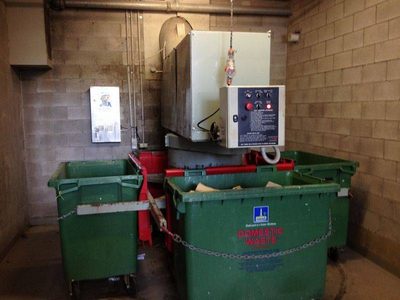 Garbage Doctor Platinum beside a waste compactor's controls, above green domestic waste bins