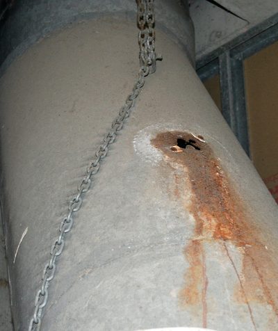 A hole eaten through a round steel waste chute by a spark-type ozone generator, rust running down below it