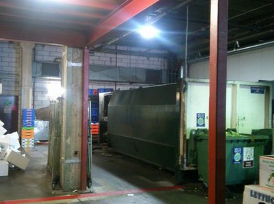 A covered loading dock with a large green waste compactor and rubbish bins