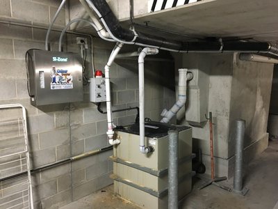 A Garbage Doctor odour control system on a block wall beside an above-ground grease arrestor, with white PVC pipework running from the machine to the grease trap
