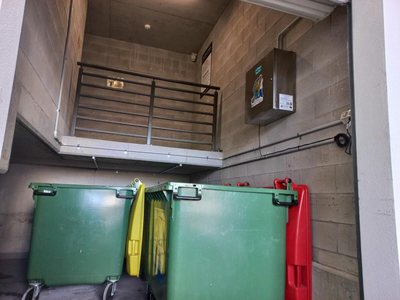 A Garbage Doctor odour control system on the block wall of a commercial bin area, above large green bins and red- and yellow-lidded bins