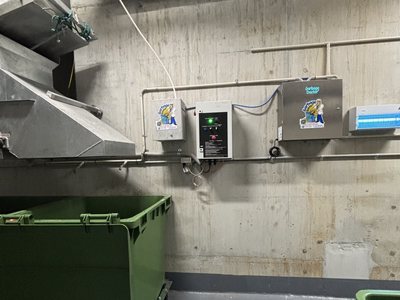 A Garbage Doctor Duo's main unit and scent tank on a concrete bin room wall, above a green bin